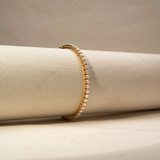 Side view of a diamond tennis bracelet in 14 KT Yellow Gold, featuring a continuous row of round brilliant diamonds in a sleek linear setting, displayed against a soft beige backdrop.