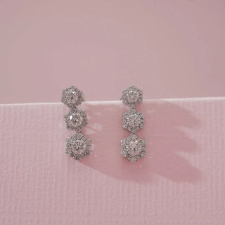 Front view of white gold hexagon triple cluster diamond drop earrings featuring round lab-grown diamonds arranged in geometric stacked formations, displayed on a soft pink textured surface.