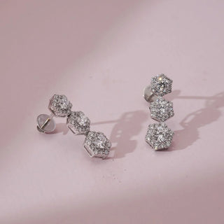 Angled view of hexagon triple cluster diamond drop earrings with round lab-grown diamonds set in stacked geometric frames, displayed on a soft pink surface with gentle studio lighting.