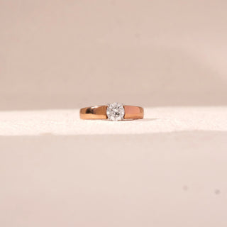 Front view of a 14KT rose gold ring featuring a round-cut diamond, set against a beige background.
