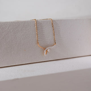 Side angled view of diamond pendant necklace in yellow gold, featuring a curved lab-grown diamond-studded design with a central baguette diamond. Set against a soft beige textured background.