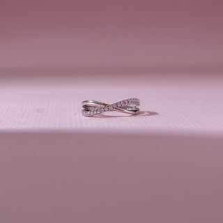 Front view of a white gold crossover diamond ring, featuring intertwined pavé-set bands with flowing curves. Captured on a soft pink backdrop, highlighting modern elegance and sparkle.
