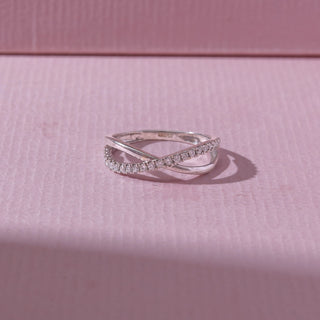 Top view of a white gold crossover diamond ring, featuring intertwined pavé-set bands with elegant curved lines. Captured on a soft pink backdrop, highlighting graceful sparkle and modern style.
