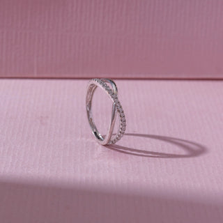 Angled view of a white gold crossover diamond ring, featuring intertwined pavé-set bands with flowing curves. Captured on a soft pink backdrop, highlighting delicate sparkle and elegance.
