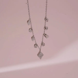 Front view of white gold diamond charm necklace with multiple round and geometric lab grown diamond drops and a central diamond-shaped pendant on a fine chain against a soft pink background.