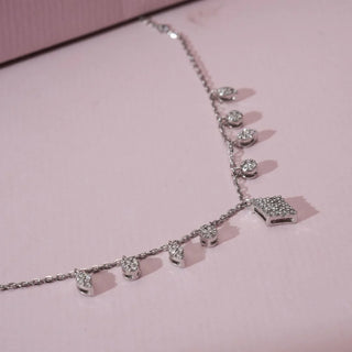 Angled view of white gold diamond charm necklace featuring multiple round lab grown diamond drops and a geometric diamond pendant resting on a soft pink background.