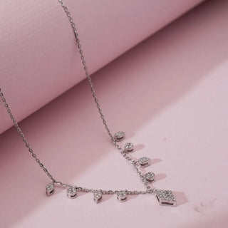 Angled close view of white gold diamond charm necklace with multiple round lab grown diamond drops and a geometric pavé diamond pendant displayed on a soft pink surface.