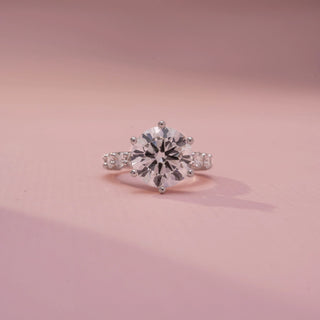 Front view of a round solitaire ring in White Gold, featuring a round-cut lab-grown diamond in a six-prong setting with diamond-accented shoulders, displayed against a soft pink backdrop.