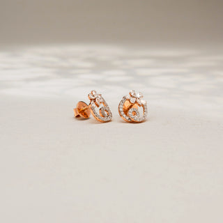 Angled view of floral diamond stud earrings in 14 KT Yellow Gold, featuring pavé-set swirling motifs with marquise and round diamond accents, displayed on a textured stone surface.
