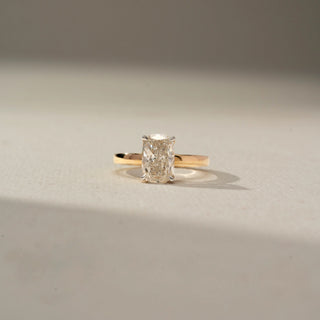 Front view of a yellow gold solitaire ring featuring an elongated radiant-cut diamond in a four-prong setting, displayed on a soft neutral surface with warm natural lighting.