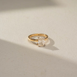 Top view of a yellow gold solitaire ring featuring an elongated radiant-cut diamond in a four-prong setting, showcased on a soft ivory surface with warm natural lighting.