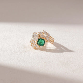 Top angled view of Multi-Stone Statement Ring in 14KT Yellow Gold, featuring an emerald-cut green gemstone, a large white diamond, and marquise accents. Set on a beige surface, it radiates bold brilliance.
