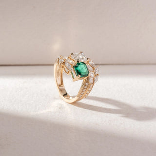 Angled view of Multi-Stone Statement Ring in 14KT Yellow Gold, featuring an emerald-cut green gemstone, a large white diamond, and marquise accents. Set on a beige surface, it reflects bold elegance and brilliance.

