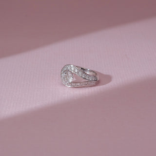 Angled view of solitaire diamond ring in white gold, featuring a round lab-grown diamond set within a curved teardrop frame lined with round diamonds. Set on a soft pink surface, it highlights fluid elegance and sparkle.