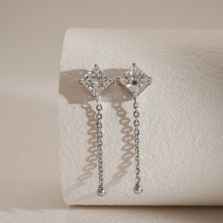 Front view of Solitaire Chain Drop Diamond Earrings against beige background. Made in 14KT hallmarked white gold, secure bombay screw closure. Set with EF colour, VVS clarity lab grown diamonds weighing 2.18 carats, solitaire 1.09 carats. Product weighs 2.94 grams, priced INR 1,00,000–3,00,000.