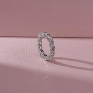Angled view of eternity band in white gold with square and rectangular diamonds in prong settings, set on a soft pink textured background, highlighting continuous design and brilliant sparkle.
