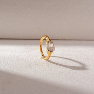 Angled view of round diamond solitaire ring in yellow gold with a smooth sculpted band, set on a soft neutral background, highlighting a classic design and gentle, radiant sparkle.
