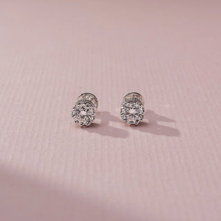 Front view of round brilliant diamond stud earrings in a white metal prong setting, displayed on a soft pink textured surface with delicate shadows and sparkling facets.