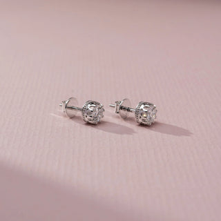 Side view of round brilliant diamond stud earrings in an intricate white metal basket setting, displayed on a soft pink textured surface with subtle shadows highlighting the profile design.