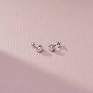 Angled view of round brilliant diamond stud earrings in ornate white metal basket settings, displayed on a soft pink textured surface with delicate shadows and sparkling details.
