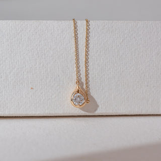 Front view of solitaire diamond pendant necklace in yellow gold, featuring a round lab-grown diamond set within a smooth teardrop frame. Set against a textured neutral backdrop, it reflects minimal elegance.