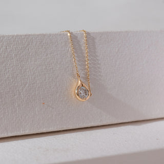 Angled view of solitaire diamond pendant necklace in yellow gold, featuring a round lab-grown diamond set within a smooth teardrop frame. Set on a textured neutral surface, it highlights soft elegance and sparkle.