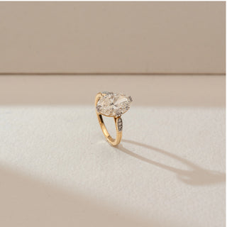 Side angled view of oval diamond ring in yellow gold, featuring a central lab-grown diamond with pavé side accents. Captured against a soft beige textured background with gentle shadows.