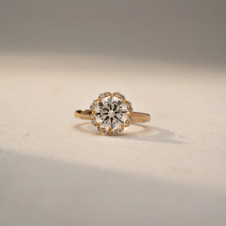 Front view of a round halo engagement ring in 14 KT Yellow Gold, featuring a round lab-grown diamond encircled by a floral-inspired halo of smaller diamonds, displayed against a beige backdrop.