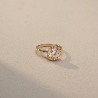 Side view of a round halo engagement ring in 14 KT Yellow Gold, featuring a round lab-grown diamond encircled by a floral-inspired halo of smaller diamonds, displayed against a beige backdrop.