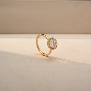 Angled view of a round halo engagement ring in 14 KT Yellow Gold, featuring a round lab-grown diamond encircled by a floral-inspired halo of smaller diamonds, displayed against a beige backdrop.