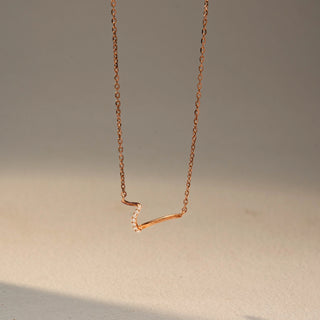 Front view of a curved diamond necklace in 14 KT Rose Gold, featuring a sleek V-shaped design accented with pavé-set lab-grown diamonds, suspended from a delicate chain against a beige backdrop.