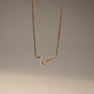 Close up view of a curved diamond necklace in 14 KT Rose Gold, featuring a sleek V-shaped design accented with pavé-set lab-grown diamonds, suspended from a delicate chain against a beige backdrop.