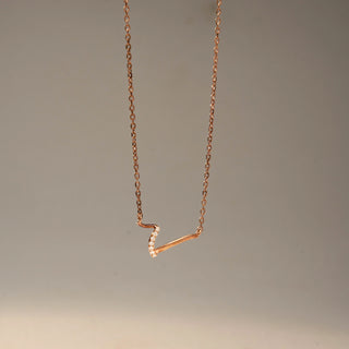 Side view of a curved diamond necklace in 14 KT Rose Gold, featuring a sleek V-shaped design accented with pavé-set lab-grown diamonds, suspended from a delicate chain against a beige backdrop.