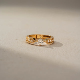 Front view of a vintage-inspired solitaire ring in 14 KT Yellow Gold, featuring a round lab-grown diamond flanked by graduated round diamonds within geometric milgrain-accented shoulders, displayed against a beige backdrop