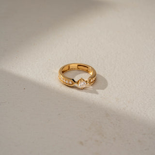 Top view of a vintage-inspired solitaire ring in 14 KT Yellow Gold, featuring a round lab-grown diamond flanked by graduated round diamonds within geometric milgrain-accented shoulders, displayed against a beige backdrop