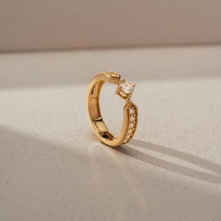 Angled view of a vintage-inspired solitaire ring in 14 KT Yellow Gold, featuring a round lab-grown diamond flanked by graduated round diamonds within geometric milgrain-accented shoulders, displayed against a beige backdrop