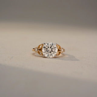 Front view of a round solitaire engagement ring in 14 KT Yellow Gold, featuring a round lab-grown diamond accented by marquise-shaped open motifs and bezel-set side diamonds, displayed against a beige backdrop.
