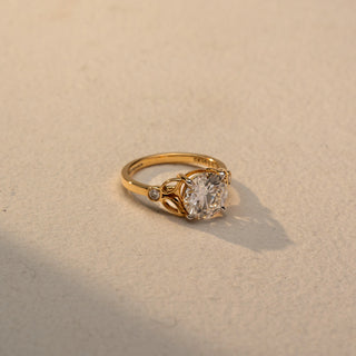 Side view of a round solitaire engagement ring in 14 KT Yellow Gold, featuring a round lab-grown diamond accented by marquise-shaped open motifs and bezel-set side diamonds, displayed against a beige backdrop.