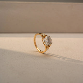 Angled view of a round solitaire engagement ring in 14 KT Yellow Gold, featuring a round lab-grown diamond accented by marquise-shaped open motifs and bezel-set side diamonds, displayed against a beige backdrop.