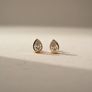 Front view of pear-shaped diamond stud earrings in yellow gold, featuring bezel-set diamonds with a sleek teardrop silhouette. Displayed against a neutral backdrop, the minimalist design sparkles elegantly.