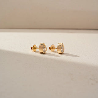Side view of pear-shaped diamond stud earrings in yellow gold, featuring bezel-set diamonds with a sleek teardrop silhouette. Displayed on a beige backdrop, the profile highlights modern craftsmanship.