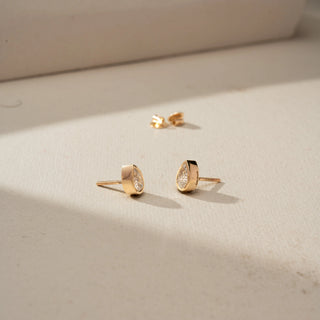 Angled view of pear-shaped diamond stud earrings in yellow gold, featuring bezel-set diamonds with a sleek teardrop silhouette. Displayed on a beige backdrop, the minimalist design shines elegantly.