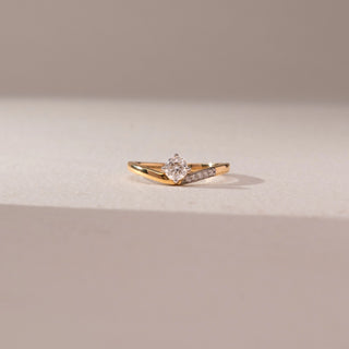 Front view of a yellow gold solitaire ring featuring a round lab-grown diamond and a pavé-set diamond accent on one shoulder. Captured against a beige backdrop, the sleek crossover design highlights modern elegance.
