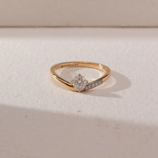 Top view of a yellow gold solitaire ring featuring a round lab-grown diamond and pavé-set accent diamonds along one shoulder. Captured against a beige backdrop, the tapered band design highlights refined elegance and sparkle.