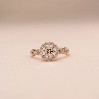 Front view of Reia’s 14 KT Yellow Gold Eternal Halo Ring. The centred round diamond with its sparkling halo sits on a diamond-studded infinity band against a soft pink surface.