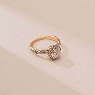 Top view of Reia’s 14 KT Yellow Gold Eternal Halo Diamond Solitaire Ring. A round diamond with a sparkling halo sits on a diamond-studded infinity band against a soft pink surface.