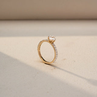 Angled view of a princess-cut solitaire engagement ring in 14 KT Yellow Gold, featuring a princess-cut lab-grown diamond set on a slim pavé diamond band, displayed against a beige backdrop.