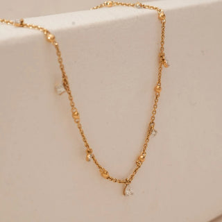 Side view of yellow gold necklace with polished bead stations and delicate dangling pear-shaped lab grown diamond accents, featuring a central pear diamond drop against a soft neutral background.