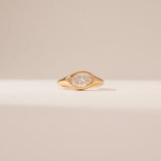 Front view of a bezel-set diamond ring in 14KT Yellow Gold, featuring an oval centre diamond and a sculpted band against a beige backdrop.