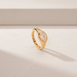 Angled view of a bezel-set diamond ring in 14KT Yellow Gold, featuring an oval centre diamond and a sculpted band against a beige backdrop.